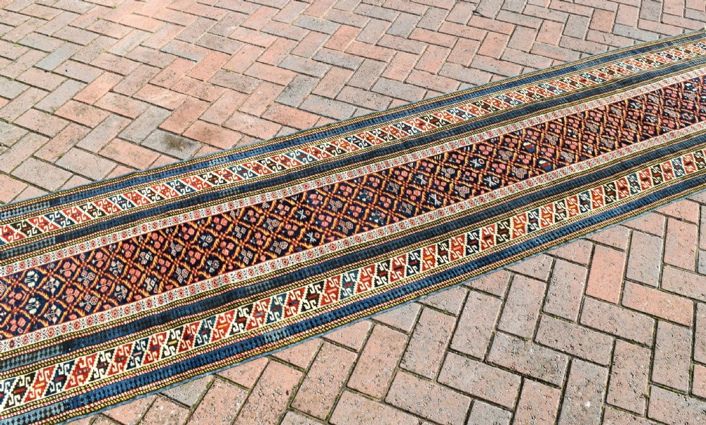 A long, narrow hand-knotted antique Caucasian Kuba runner rug featuring a navy blue geometric diamond lattice pattern framed by intricate multi-coloured borders. View IV
