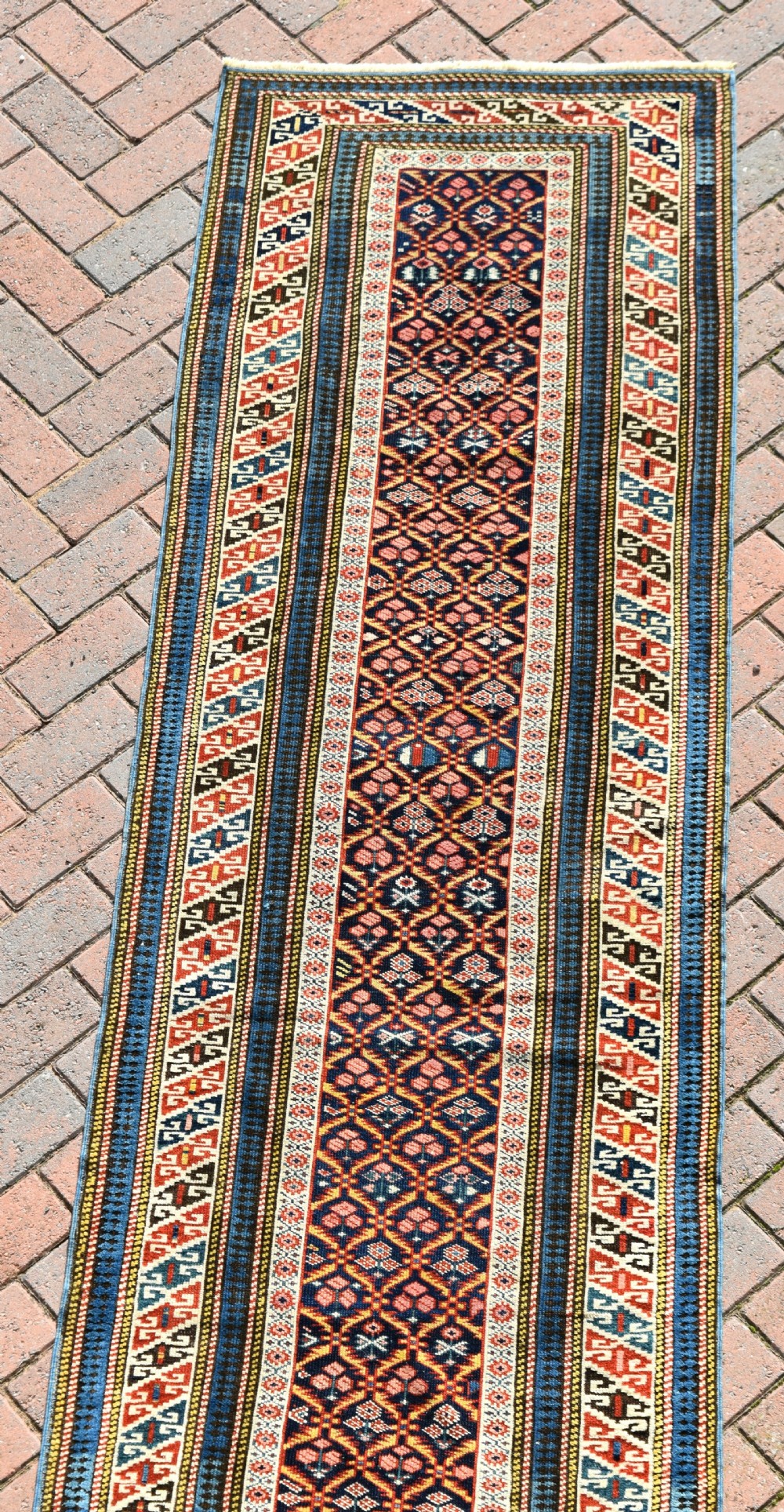 A long, narrow hand-knotted antique Caucasian Kuba runner rug featuring a navy blue geometric diamond lattice pattern framed by intricate multi-coloured borders. View VII
