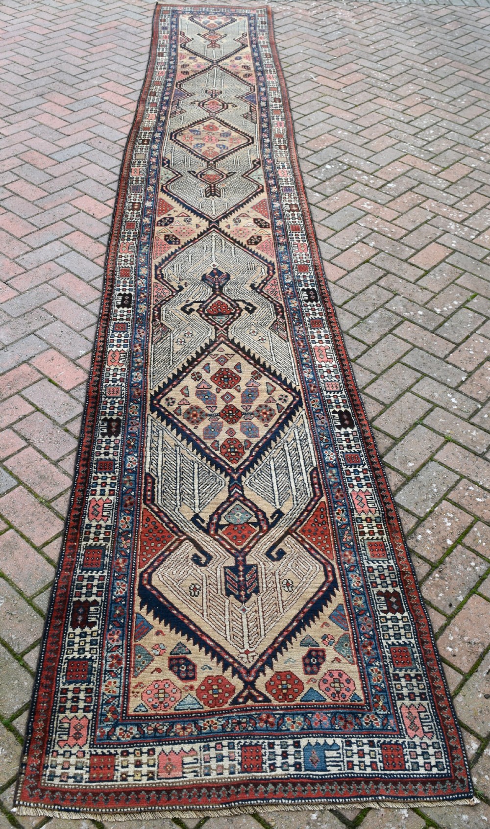 A long, narrow antique hand-knotted Serab wool runner rug featuring repeating geometric diamond medallions in beige, terracotta red, pink, and navy blue tones. View II