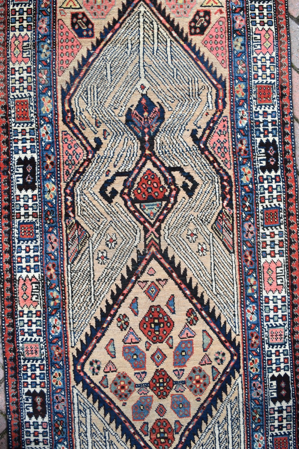A long, narrow antique hand-knotted Serab wool runner rug featuring repeating geometric diamond medallions in beige, terracotta red, pink, and navy blue tones. View IV