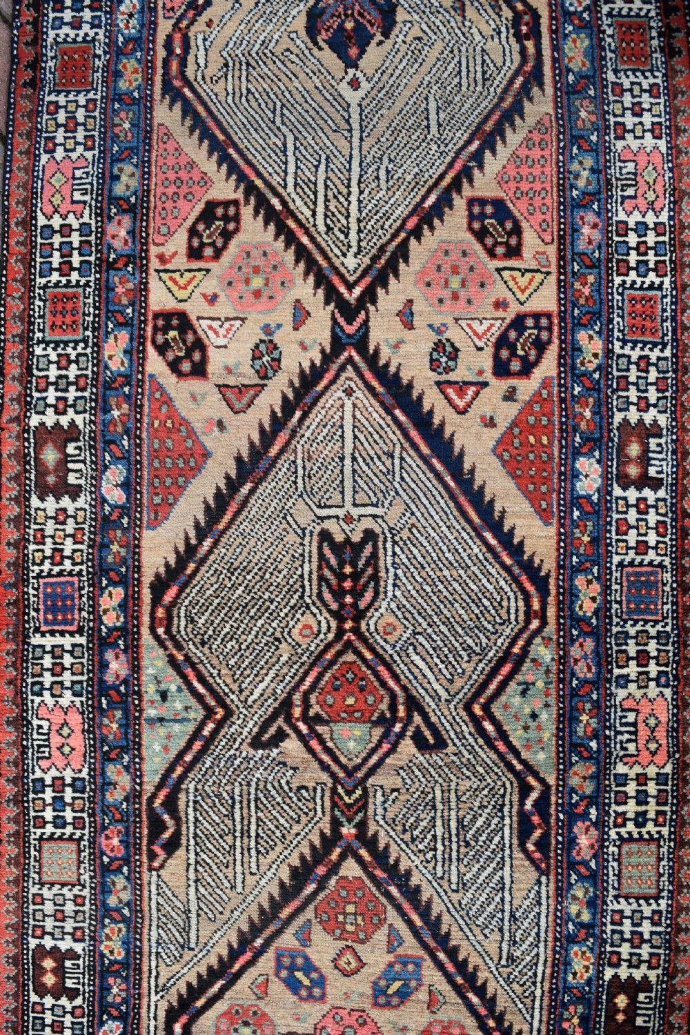 A long, narrow antique hand-knotted Serab wool runner rug featuring repeating geometric diamond medallions in beige, terracotta red, pink, and navy blue tones. View V