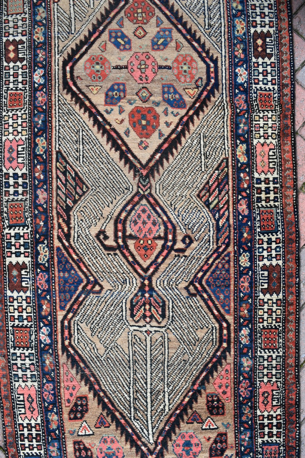 A long, narrow antique hand-knotted Serab wool runner rug featuring repeating geometric diamond medallions in beige, terracotta red, pink, and navy blue tones. View VI