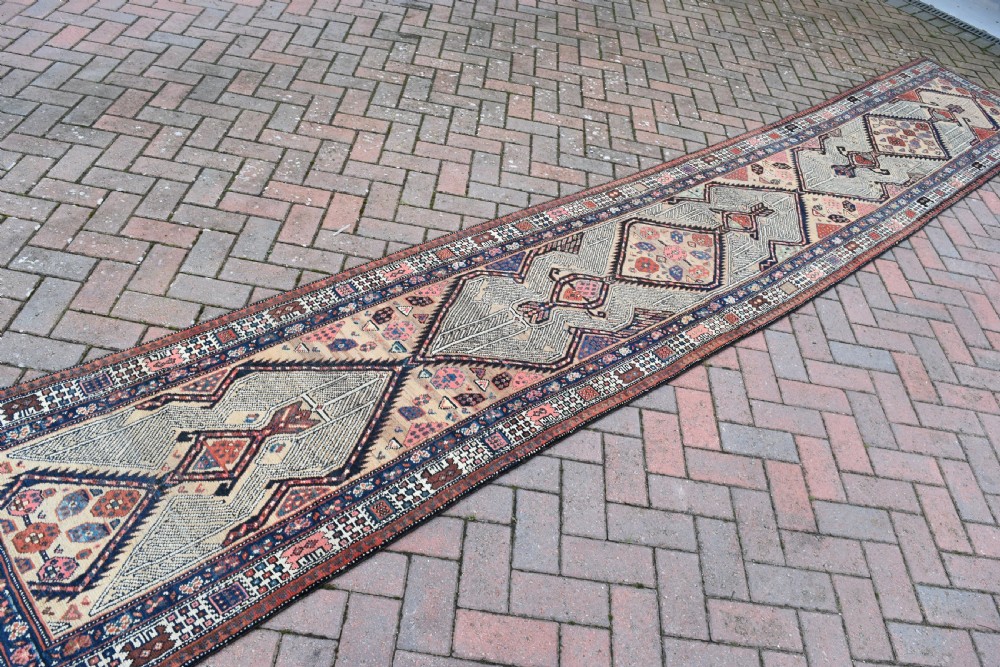 A long, narrow antique hand-knotted Serab wool runner rug featuring repeating geometric diamond medallions in beige, terracotta red, pink, and navy blue tones. View VIII