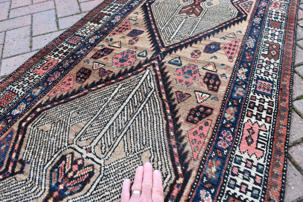 A long, narrow antique hand-knotted Serab wool runner rug featuring repeating geometric diamond medallions in beige, terracotta red, pink, and navy blue tones. View IX