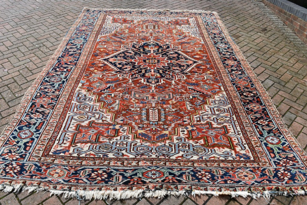 A large hand-knotted antique Heriz wool rug featuring a central geometric medallion in navy blue set against a terracotta red field with intricate corner spandrels.