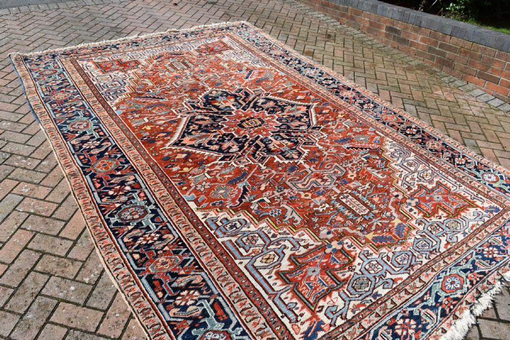 A large hand-knotted antique Heriz wool rug featuring a central geometric medallion in navy blue set against a terracotta red field with intricate corner spandrels. View III