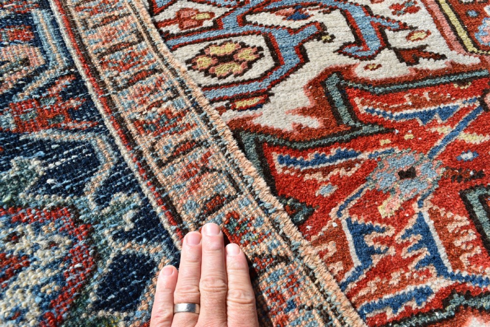 A large hand-knotted antique Heriz wool rug featuring a central geometric medallion in navy blue set against a terracotta red field with intricate corner spandrels. View IV