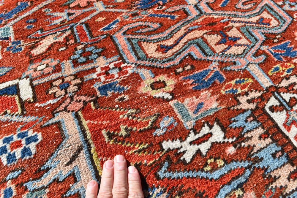 A large hand-knotted antique Heriz wool rug featuring a central geometric medallion in navy blue set against a terracotta red field with intricate corner spandrels. View V