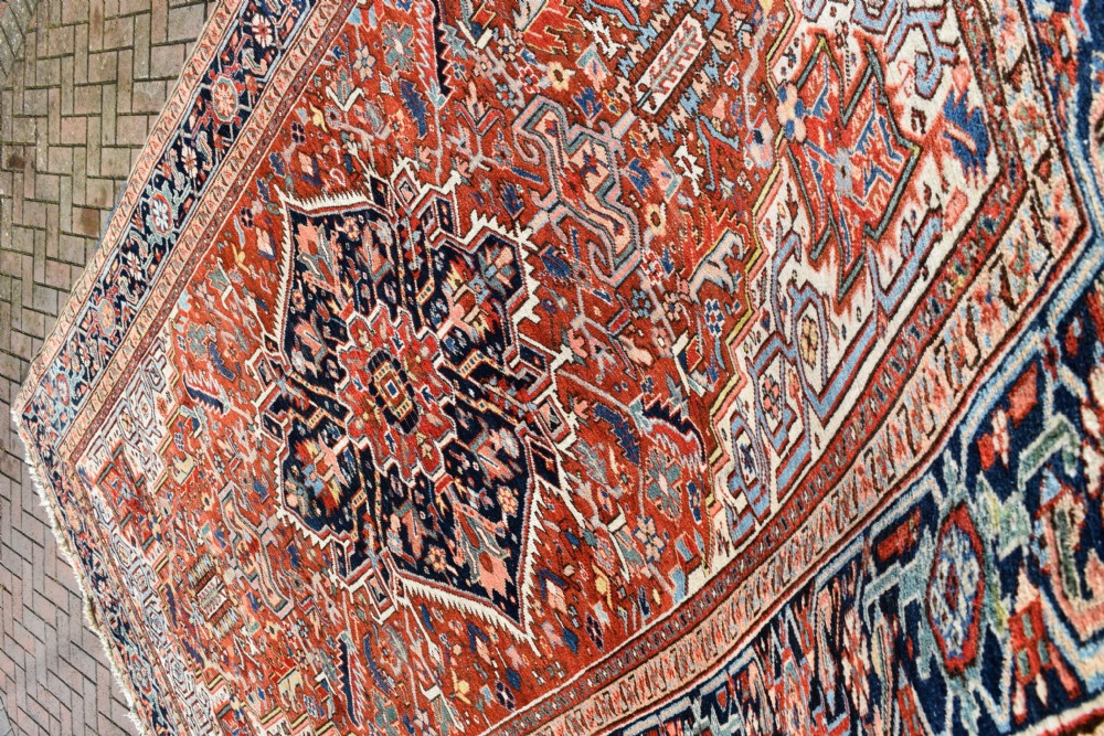 A large hand-knotted antique Heriz wool rug featuring a central geometric medallion in navy blue set against a terracotta red field with intricate corner spandrels. View VI