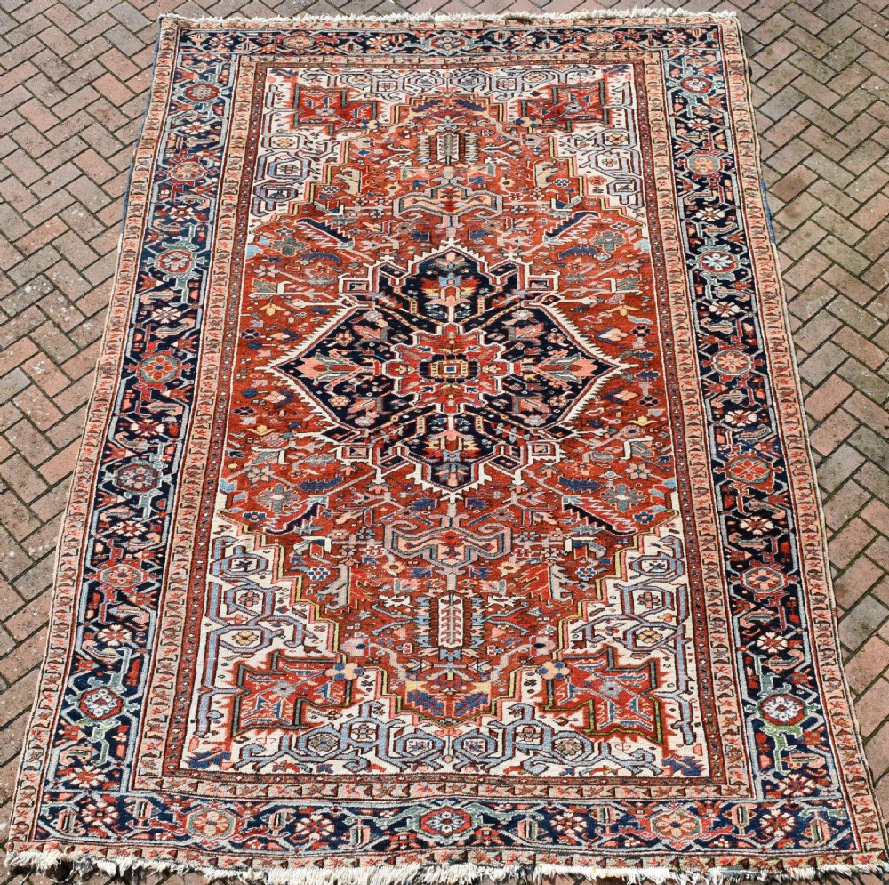 A large hand-knotted antique Heriz wool rug featuring a central geometric medallion in navy blue set against a terracotta red field with intricate corner spandrels. View VII