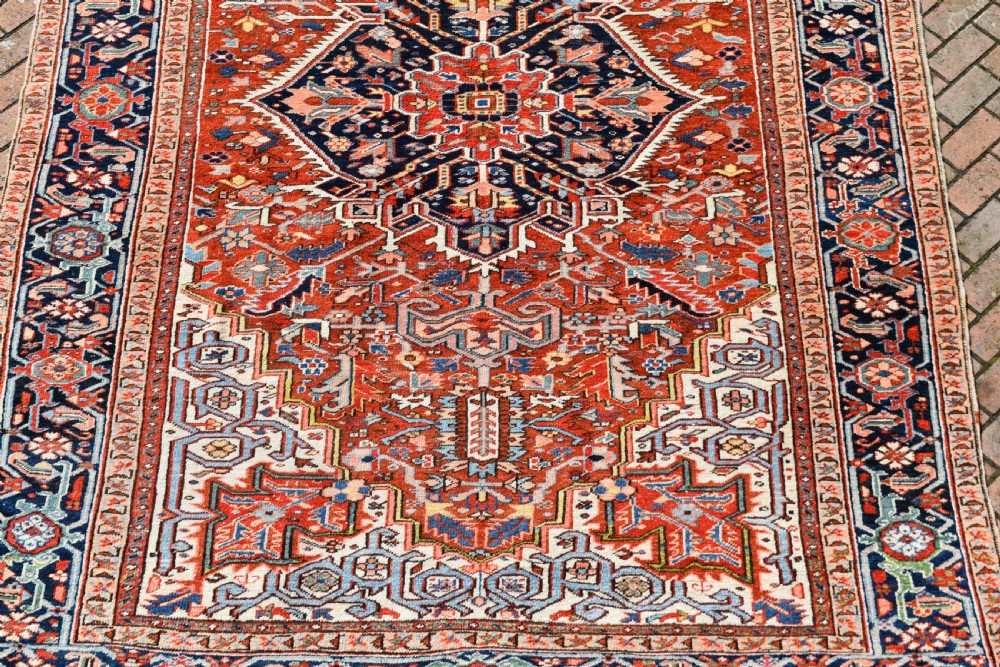 A large hand-knotted antique Heriz wool rug featuring a central geometric medallion in navy blue set against a terracotta red field with intricate corner spandrels. View VIII