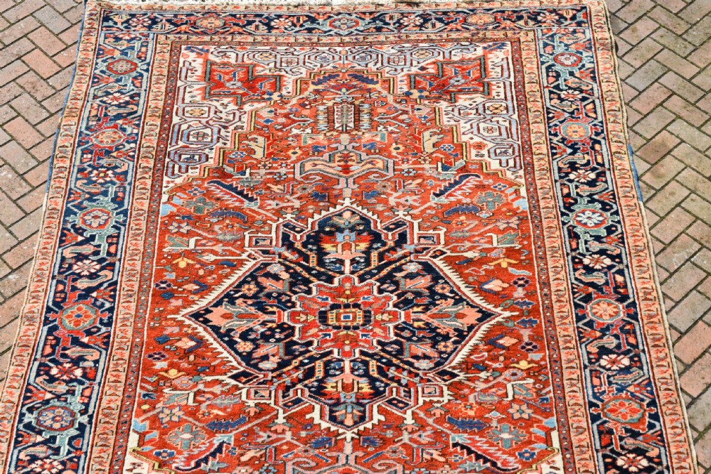 A large hand-knotted antique Heriz wool rug featuring a central geometric medallion in navy blue set against a terracotta red field with intricate corner spandrels. View IX