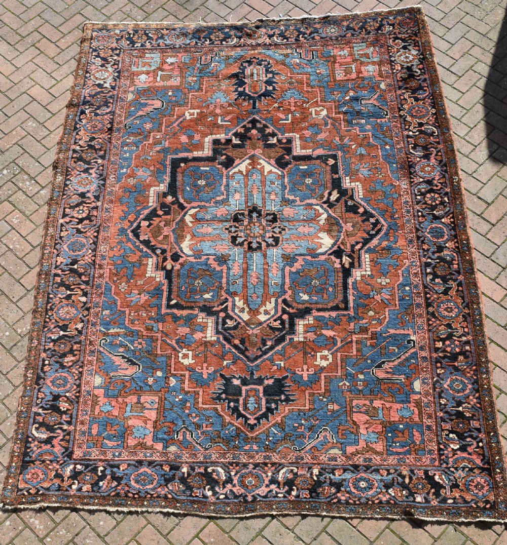 An antique hand-knotted Persian Heriz wool carpet featuring a large sky blue geometric medallion on a terracotta ground surrounded by a dark navy ornate border.