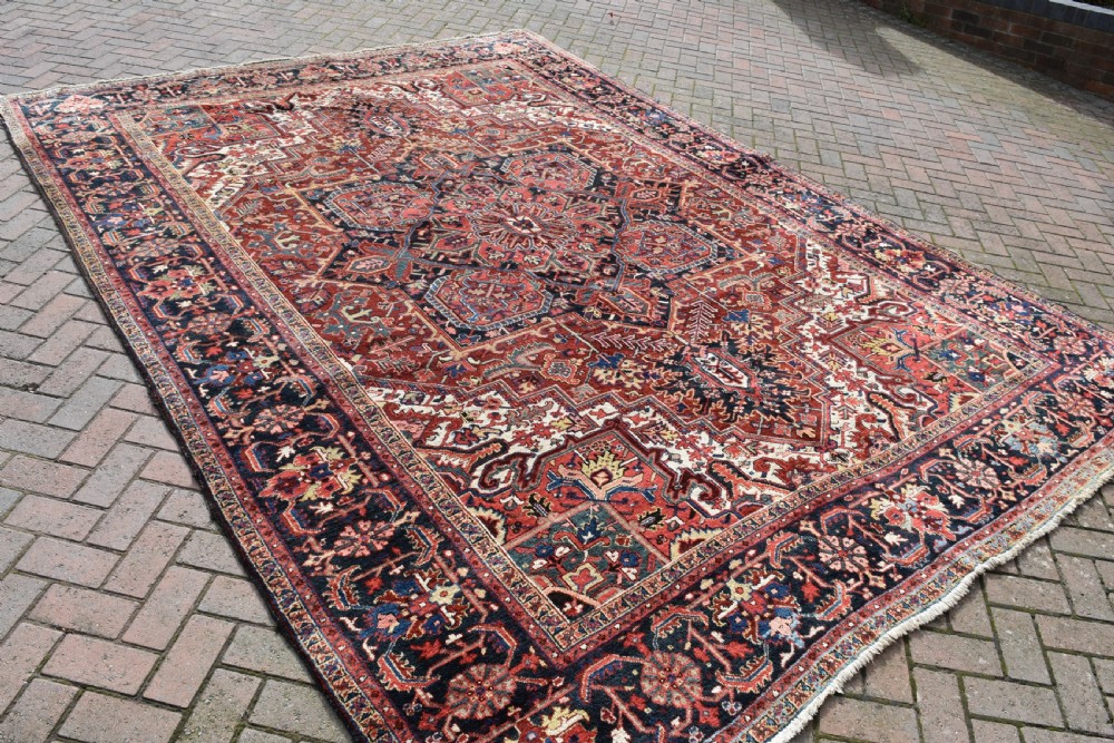 A large hand-knotted Persian Heriz wool carpet featuring a prominent central geometric medallion framed by angular corner spandrels and a multi-tiered decorative border in terracotta, navy, and cream. View III