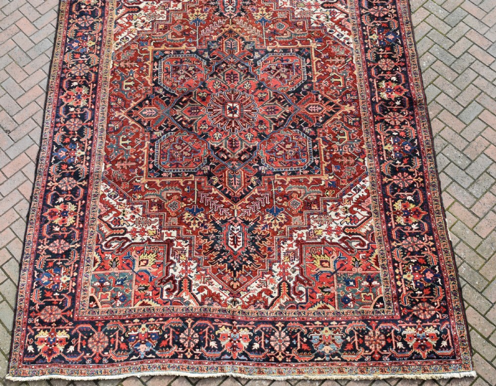 A large hand-knotted Persian Heriz wool carpet featuring a prominent central geometric medallion framed by angular corner spandrels and a multi-tiered decorative border in terracotta, navy, and cream. View V