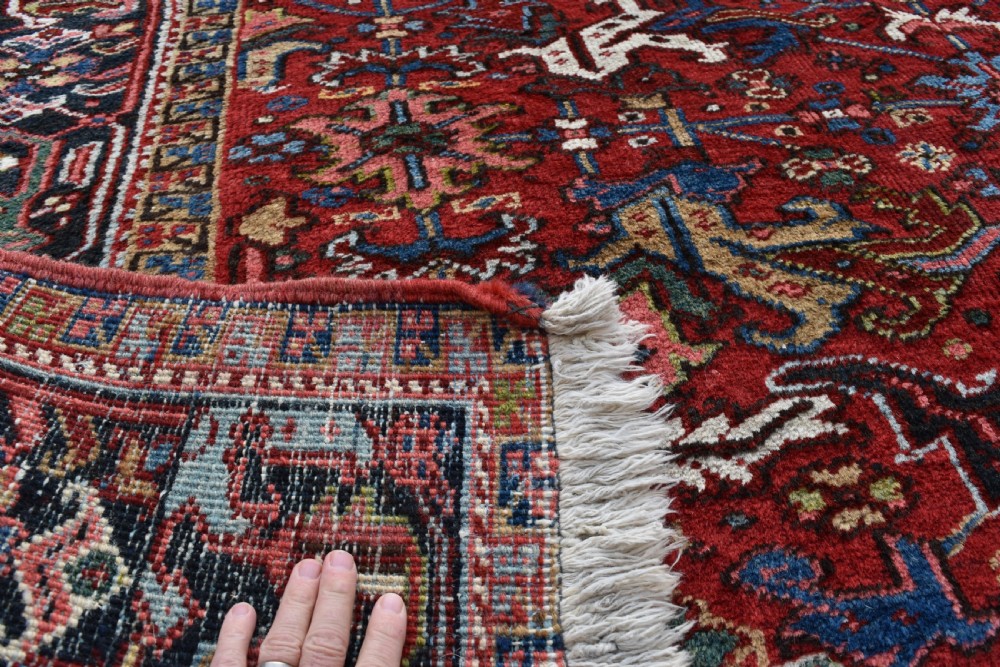 A hand-knotted Persian Heriz wool rug featuring a rich red central field with geometric patterns and a dark navy blue border. View VII