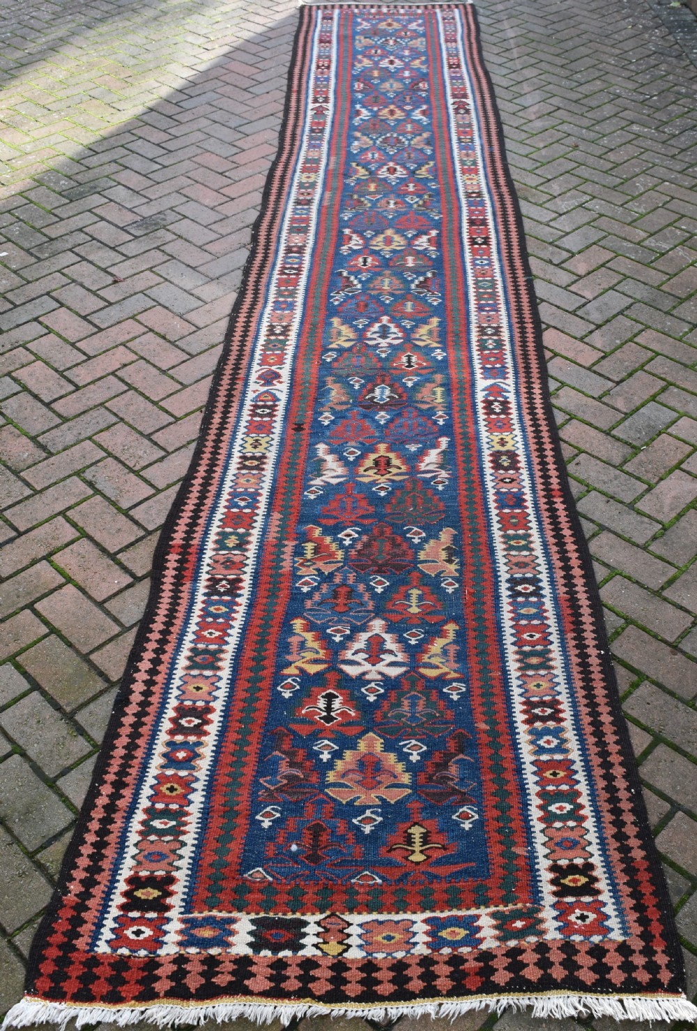 An extra long, hand-woven wool kilim runner rug featuring geometric tribal patterns in navy blue, red, cream, and ochre with fringed ends.