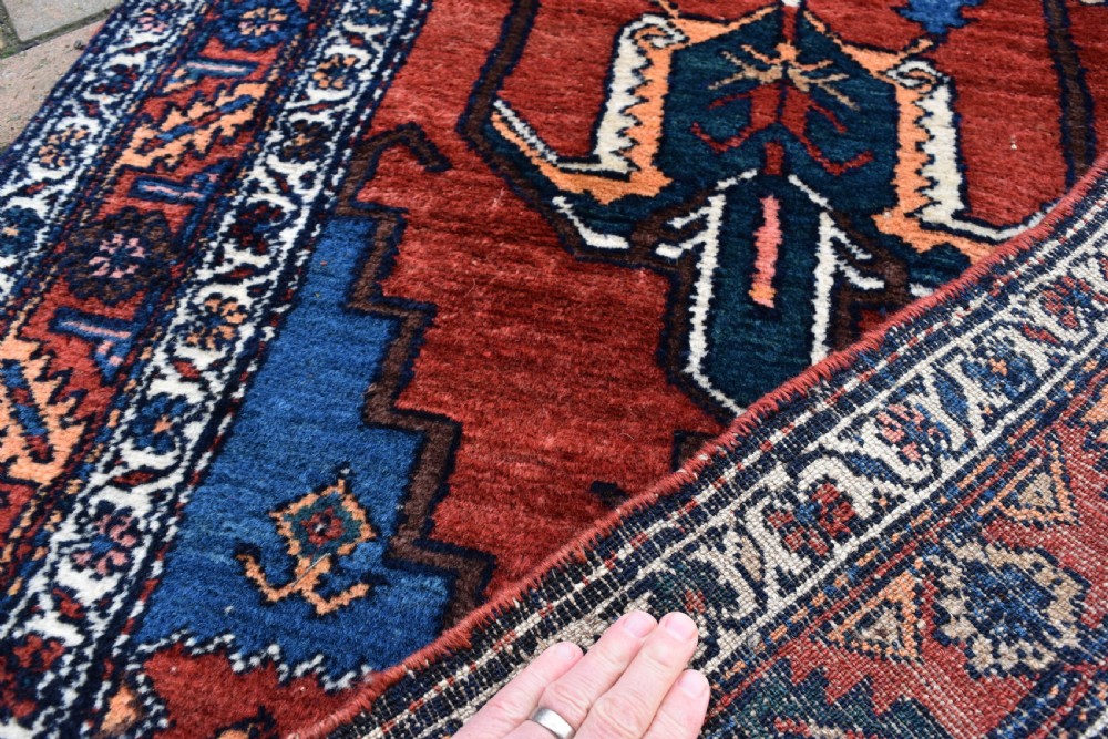 A long, hand-knotted antique Persian wool hall runner featuring geometric blue medallions along a terracotta red field with decorative borders. View IV