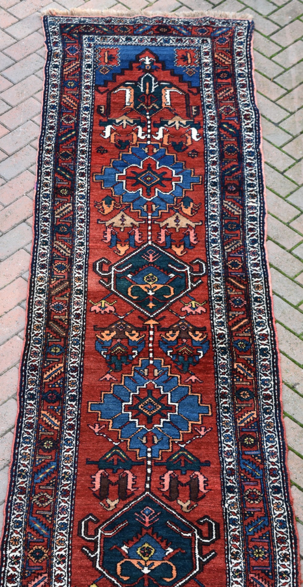 A long, hand-knotted antique Persian wool hall runner featuring geometric blue medallions along a terracotta red field with decorative borders. View V