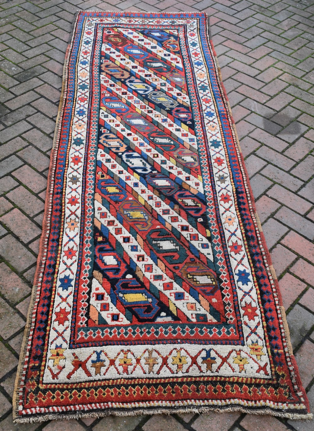 An antique Caucasian Gendje hand-knotted wool runner rug featuring diagonal geometric bands in red, navy blue, yellow, and cream enclosed by a star-motif border.