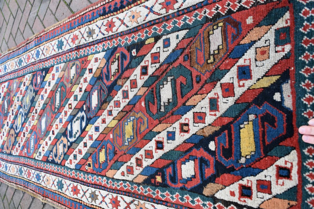 An antique Caucasian Gendje hand-knotted wool runner rug featuring diagonal geometric bands in red, navy blue, yellow, and cream enclosed by a star-motif border. View IV