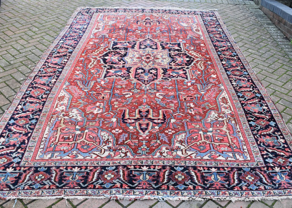 A large hand-knotted Persian Heriz wool carpet featuring a central geometric medallion surrounded by intricate angular motifs in terracotta red, navy blue, and light blue tones.