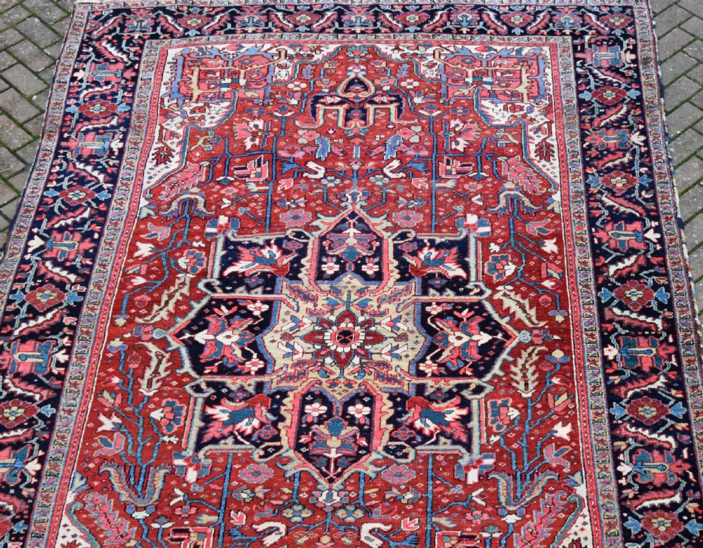 A large hand-knotted Persian Heriz wool carpet featuring a central geometric medallion surrounded by intricate angular motifs in terracotta red, navy blue, and light blue tones. View II
