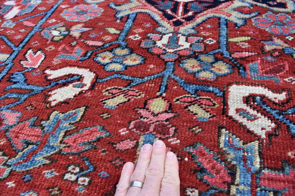 A large hand-knotted Persian Heriz wool carpet featuring a central geometric medallion surrounded by intricate angular motifs in terracotta red, navy blue, and light blue tones. View III