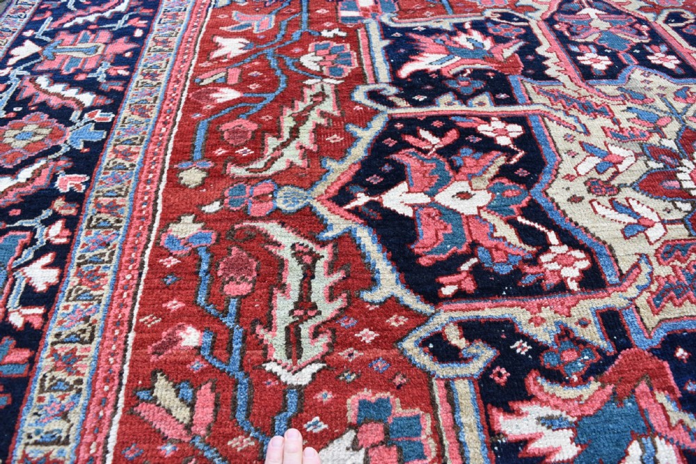 A large hand-knotted Persian Heriz wool carpet featuring a central geometric medallion surrounded by intricate angular motifs in terracotta red, navy blue, and light blue tones. View V