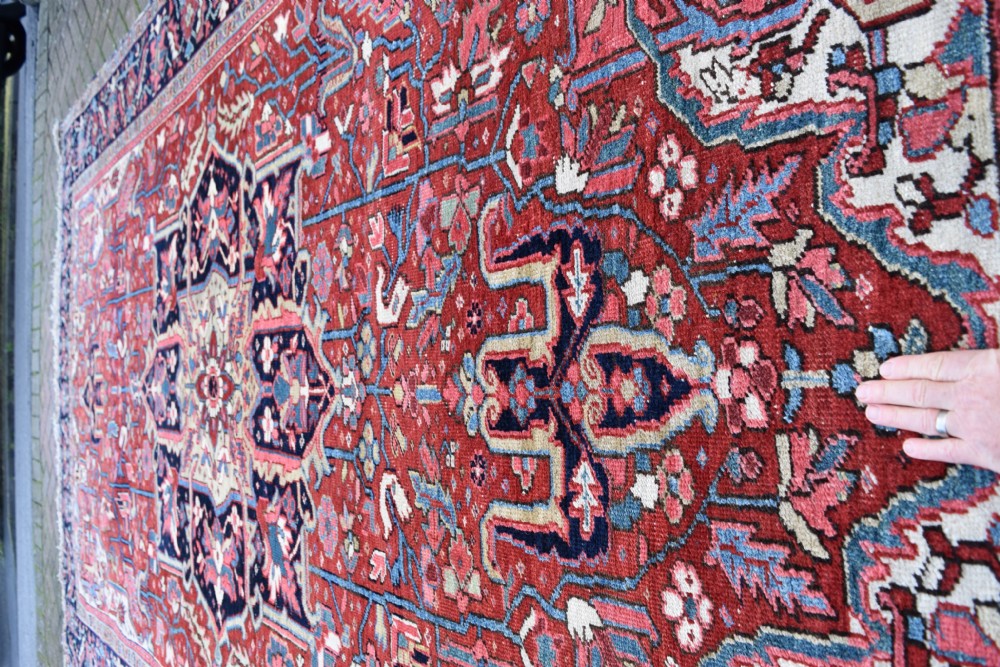 A large hand-knotted Persian Heriz wool carpet featuring a central geometric medallion surrounded by intricate angular motifs in terracotta red, navy blue, and light blue tones. View VI