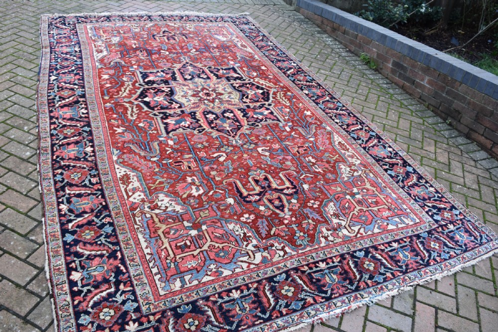 A large hand-knotted Persian Heriz wool carpet featuring a central geometric medallion surrounded by intricate angular motifs in terracotta red, navy blue, and light blue tones. View VII