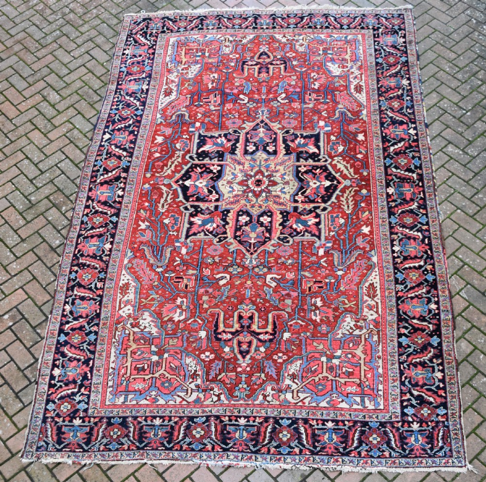 A large hand-knotted Persian Heriz wool carpet featuring a central geometric medallion surrounded by intricate angular motifs in terracotta red, navy blue, and light blue tones. View IX