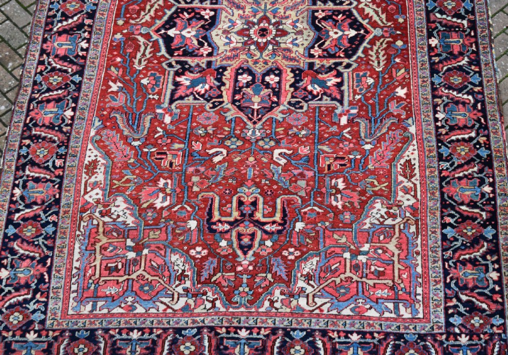 A large hand-knotted Persian Heriz wool carpet featuring a central geometric medallion surrounded by intricate angular motifs in terracotta red, navy blue, and light blue tones. View X