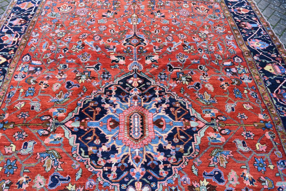 A large hand-knotted Heriz rug with a terracotta red field, central navy star medallion, and dark blue patterned border. View IV