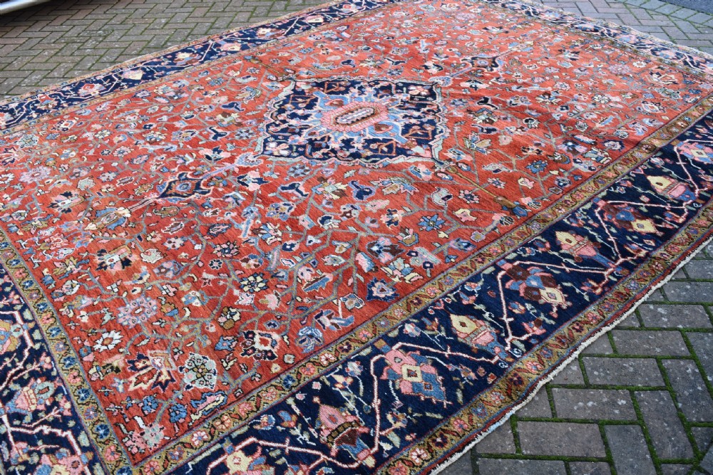 A large hand-knotted Heriz rug with a terracotta red field, central navy star medallion, and dark blue patterned border. View IX