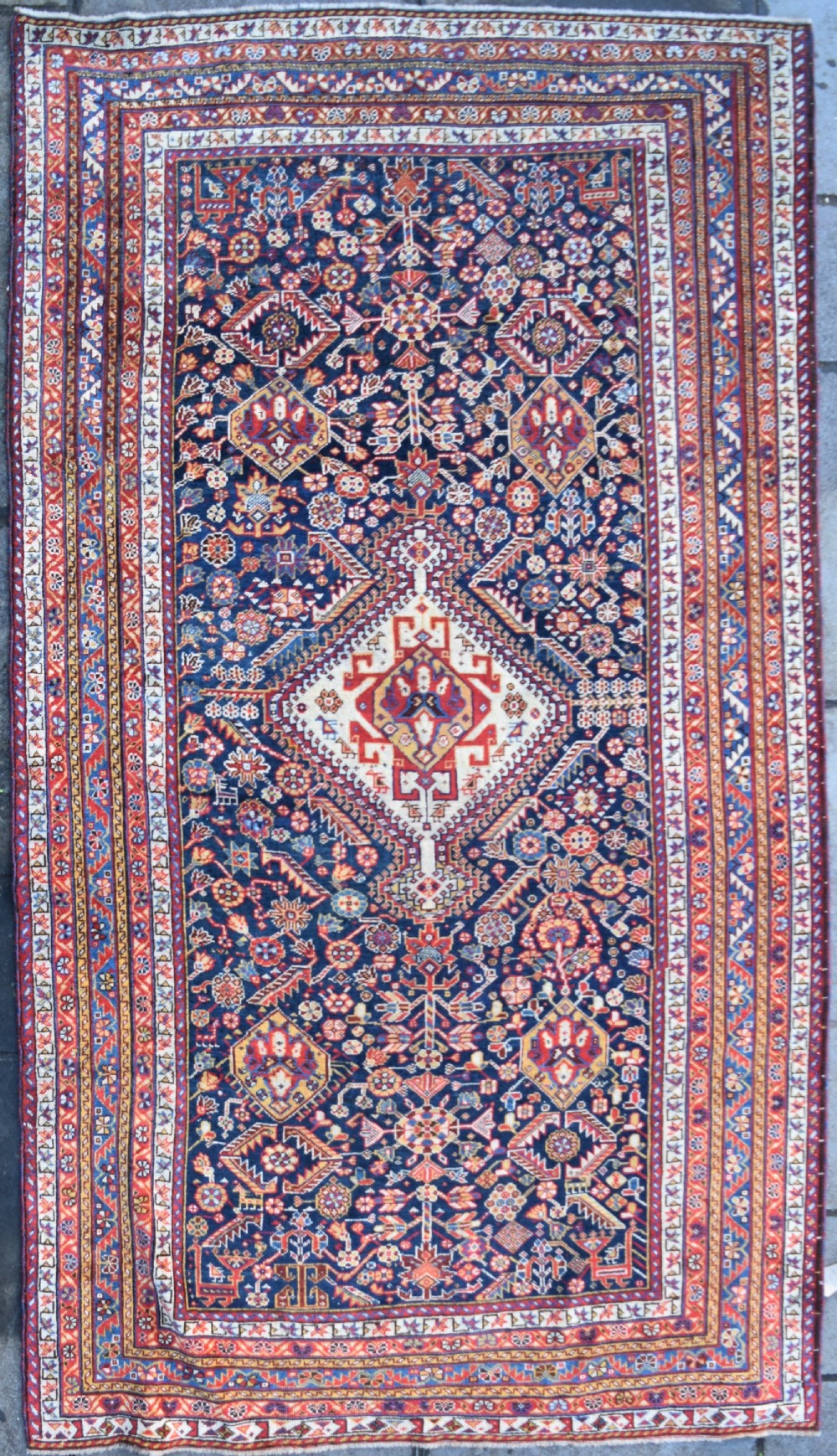 An antique hand-knotted Qashqai wool carpet with a dark navy blue field, central cream and red diamond medallion, and dense geometric border patterns.