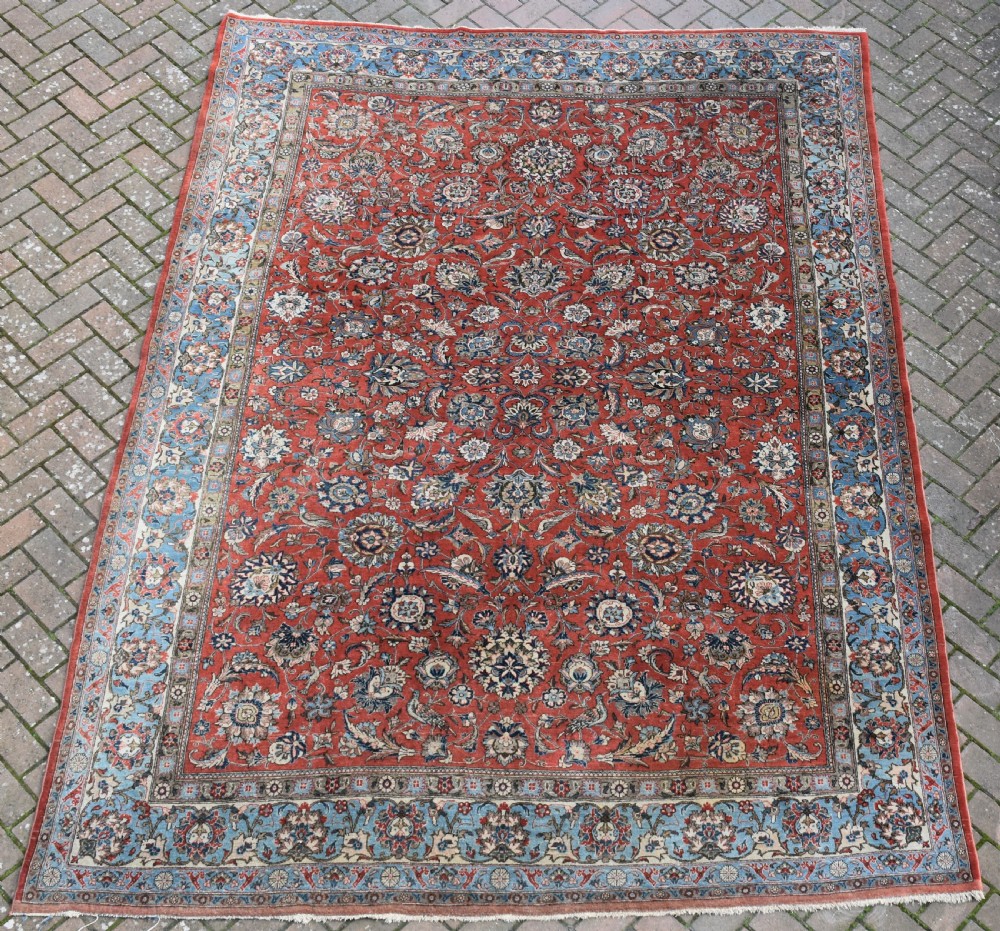 A large hand-knotted Persian Qum carpet featuring a detailed rust-red floral field framed by a wide light-blue decorative border.