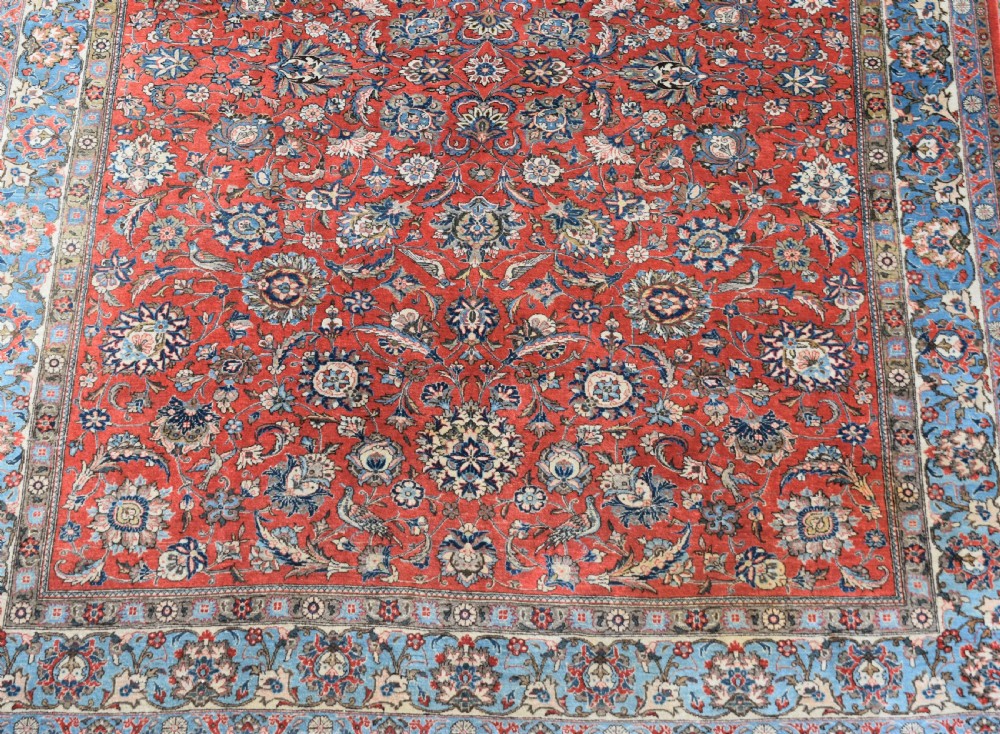 A large hand-knotted Persian Qum carpet featuring a detailed rust-red floral field framed by a wide light-blue decorative border. View II