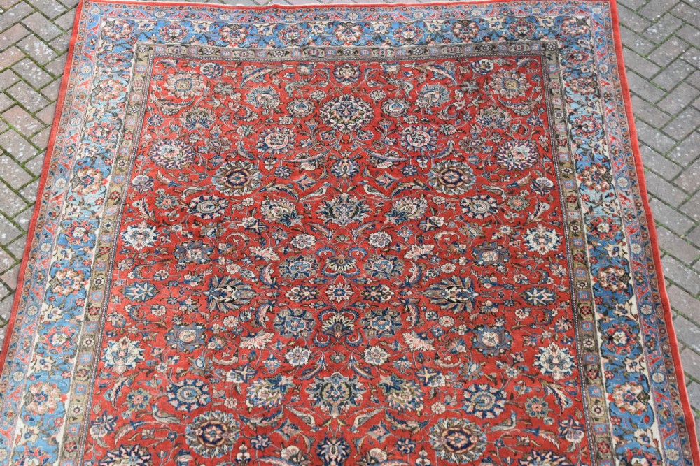 A large hand-knotted Persian Qum carpet featuring a detailed rust-red floral field framed by a wide light-blue decorative border. View III