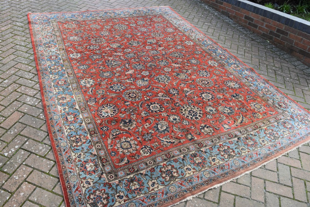 A large hand-knotted Persian Qum carpet featuring a detailed rust-red floral field framed by a wide light-blue decorative border. View IV