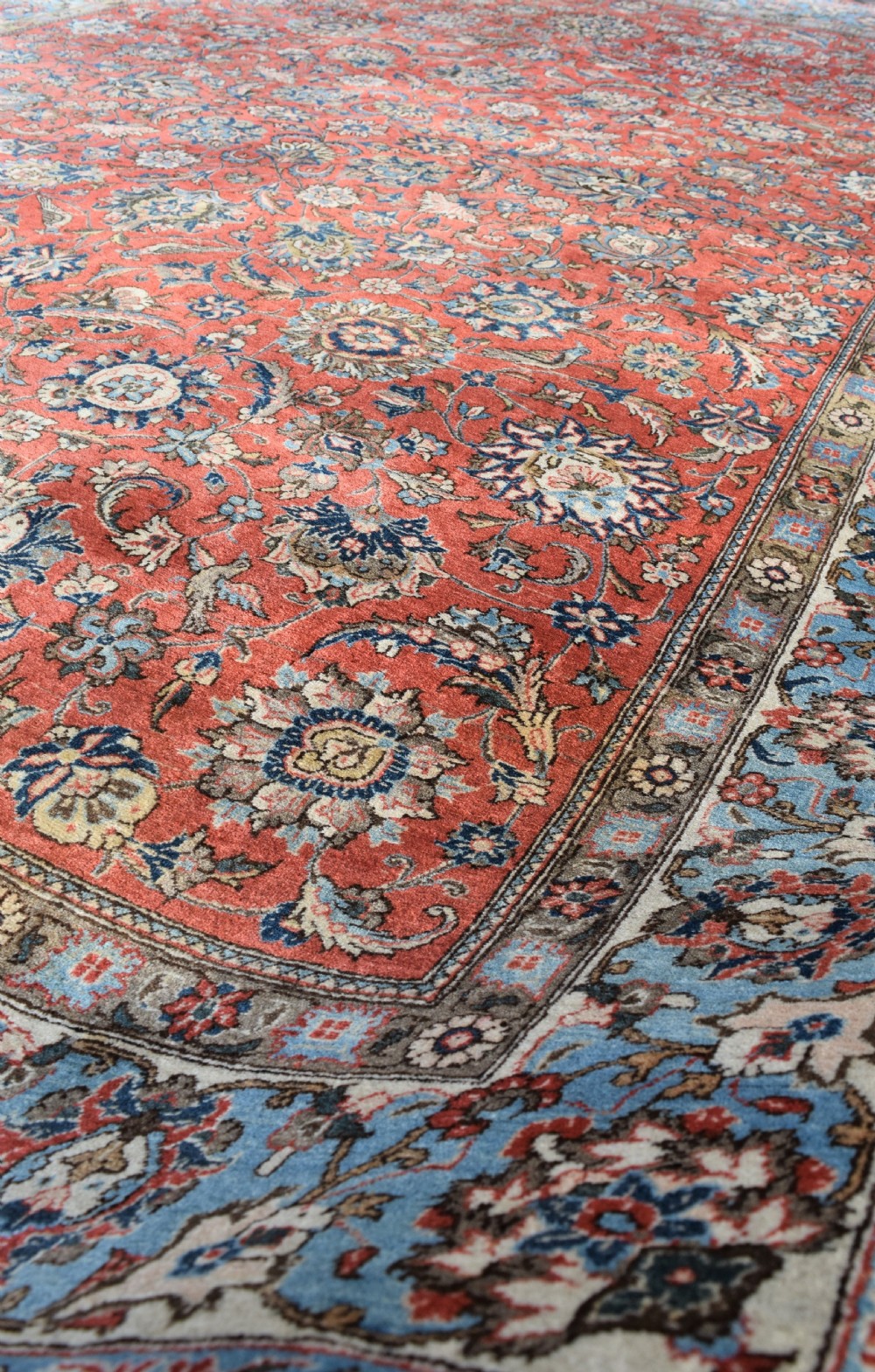 A large hand-knotted Persian Qum carpet featuring a detailed rust-red floral field framed by a wide light-blue decorative border. View VI