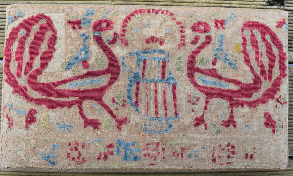 A rectangular mounted antique textile fragment featuring red silk embroidery depicting two confronting peacocks beside a central jug on a natural linen ground.