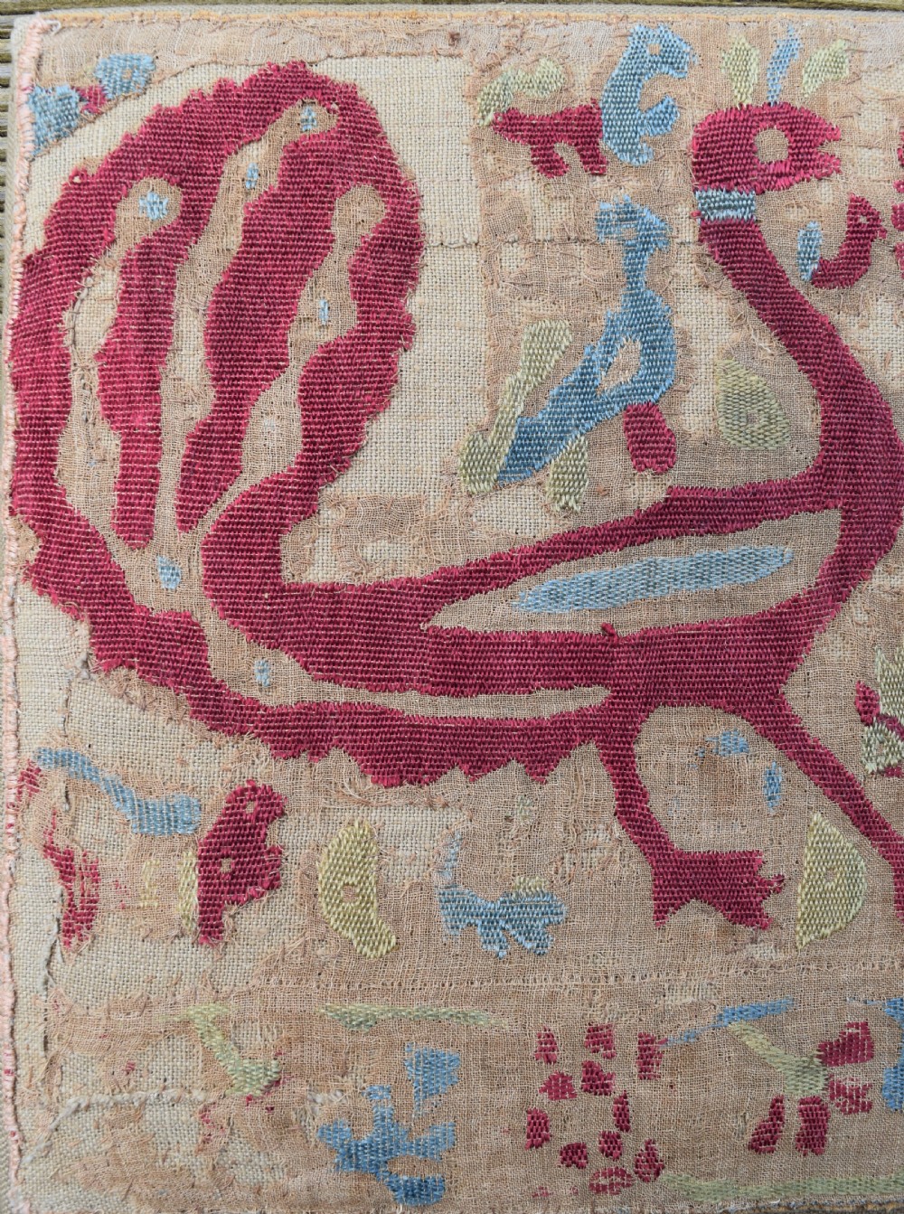 A rectangular mounted antique textile fragment featuring red silk embroidery depicting two confronting peacocks beside a central jug on a natural linen ground. View II