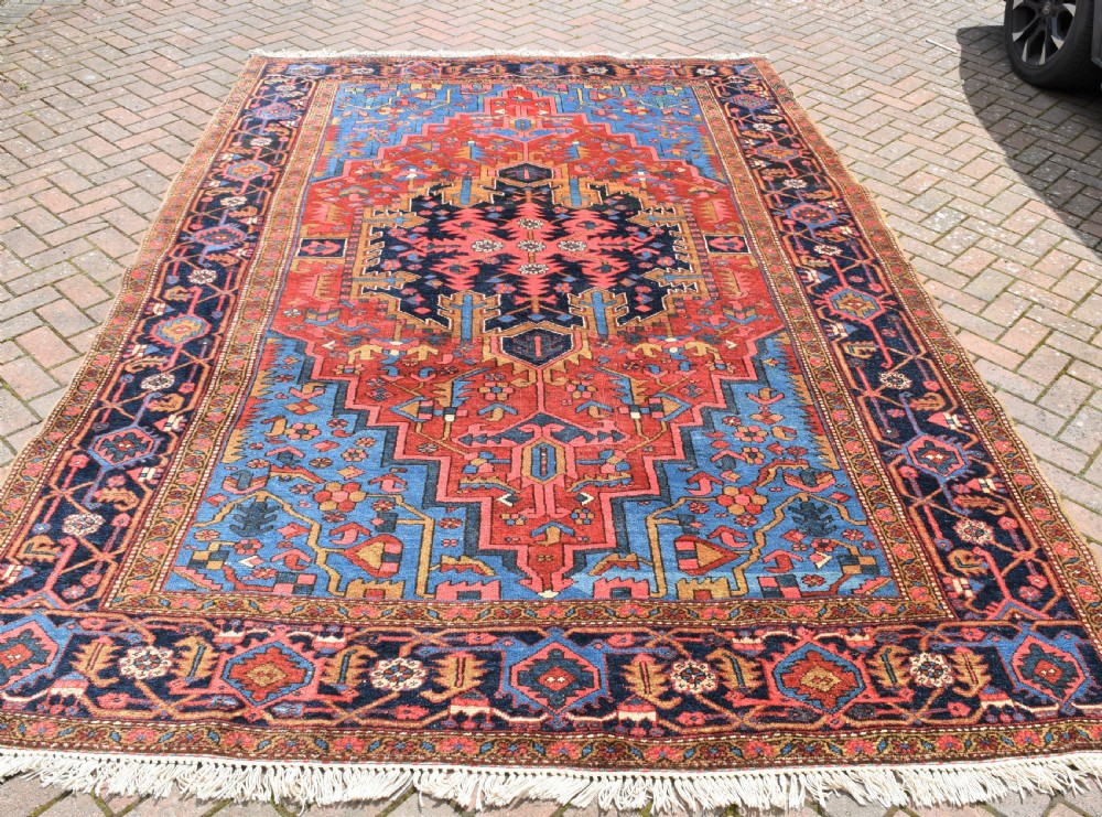 A large antique Persian Heriz wool carpet featuring a bold geometric central medallion, step-patterned spandrels in sky blue, and multi-layered decorative borders in terracotta, navy, and cream.