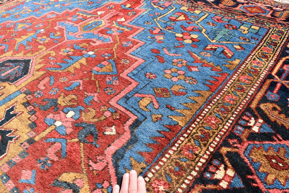 A large antique Persian Heriz wool carpet featuring a bold geometric central medallion, step-patterned spandrels in sky blue, and multi-layered decorative borders in terracotta, navy, and cream. View III
