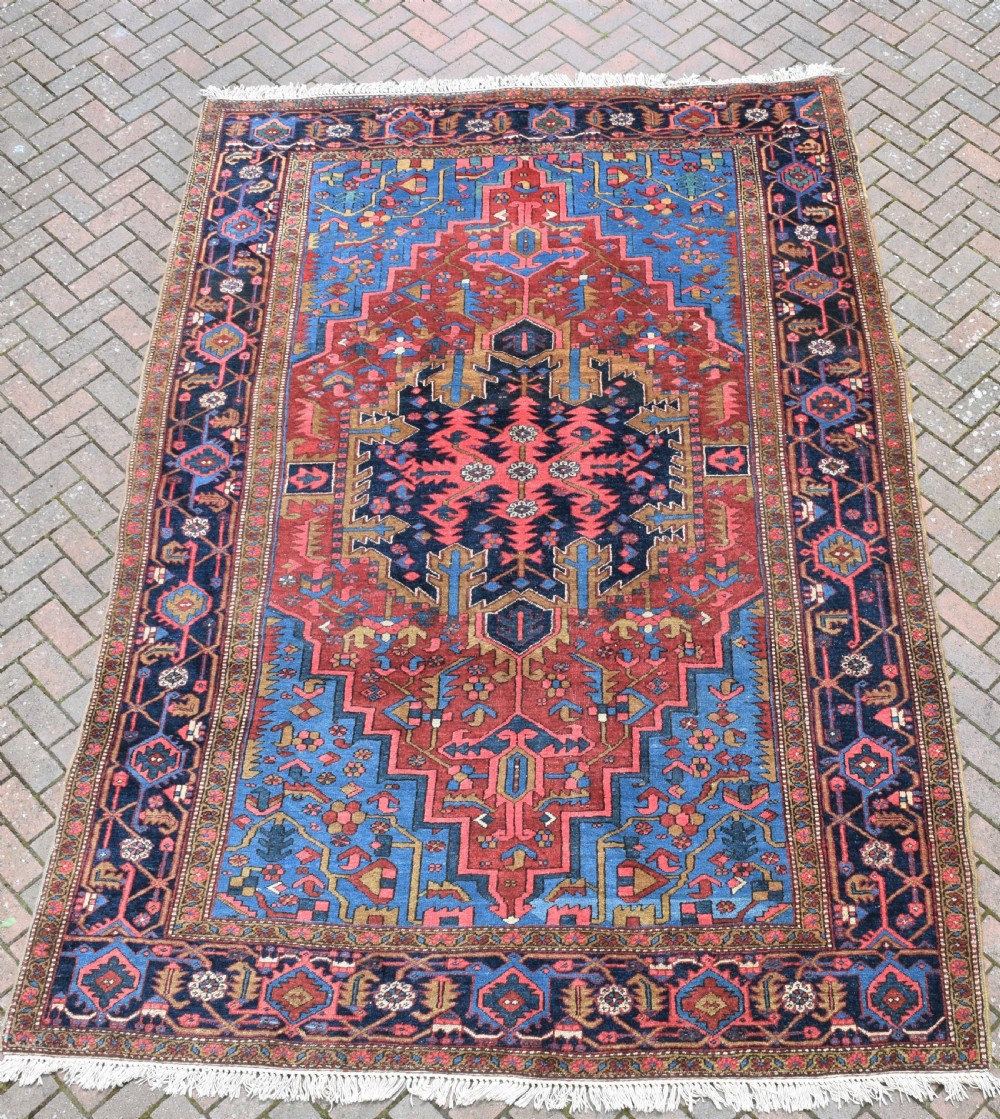 A large antique Persian Heriz wool carpet featuring a bold geometric central medallion, step-patterned spandrels in sky blue, and multi-layered decorative borders in terracotta, navy, and cream. View IV