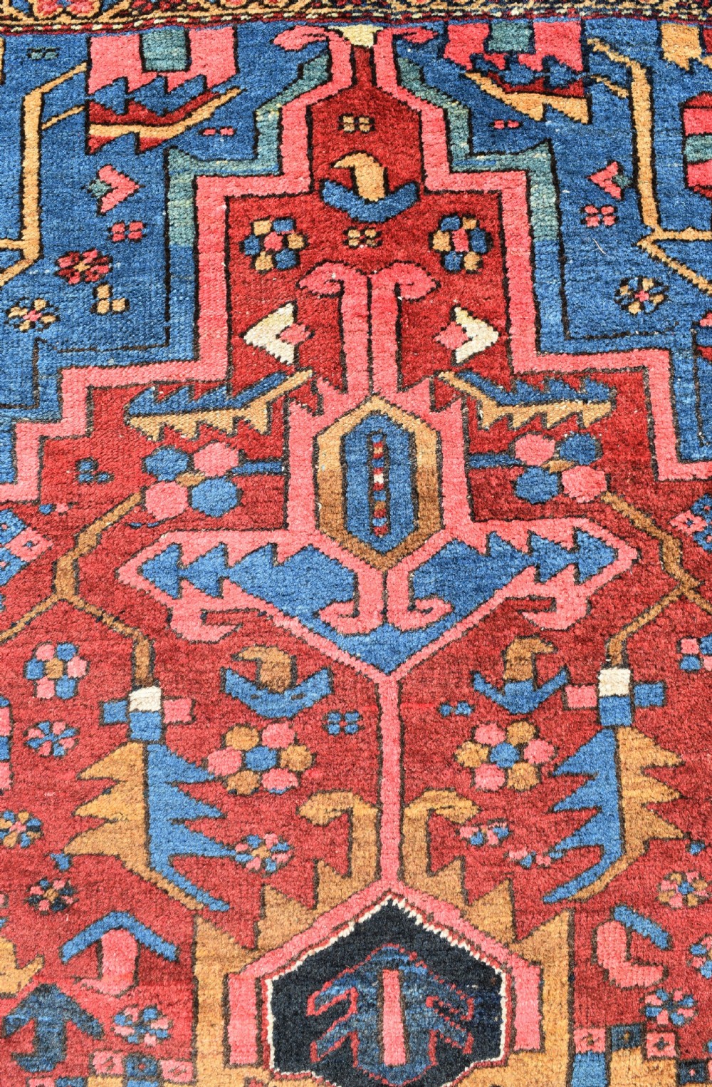 A large antique Persian Heriz wool carpet featuring a bold geometric central medallion, step-patterned spandrels in sky blue, and multi-layered decorative borders in terracotta, navy, and cream. View V