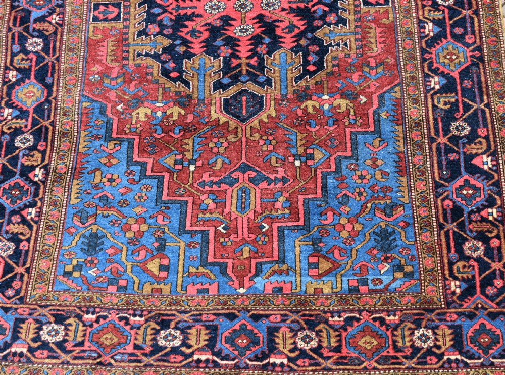 A large antique Persian Heriz wool carpet featuring a bold geometric central medallion, step-patterned spandrels in sky blue, and multi-layered decorative borders in terracotta, navy, and cream. View VI
