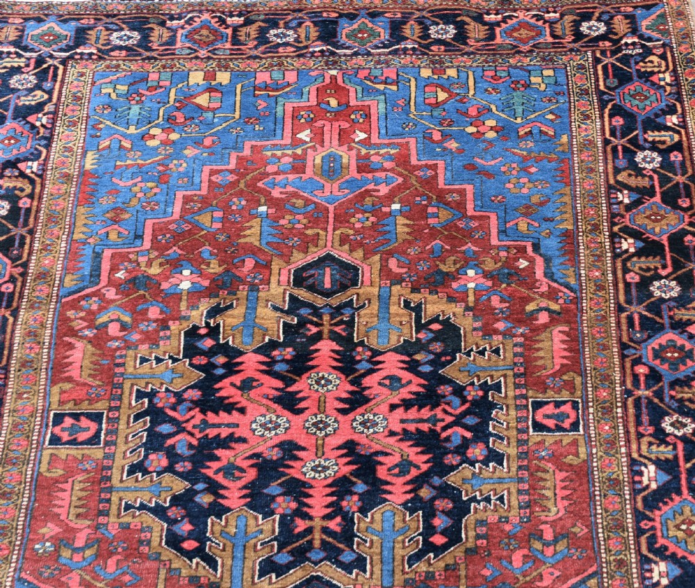 A large antique Persian Heriz wool carpet featuring a bold geometric central medallion, step-patterned spandrels in sky blue, and multi-layered decorative borders in terracotta, navy, and cream. View VII