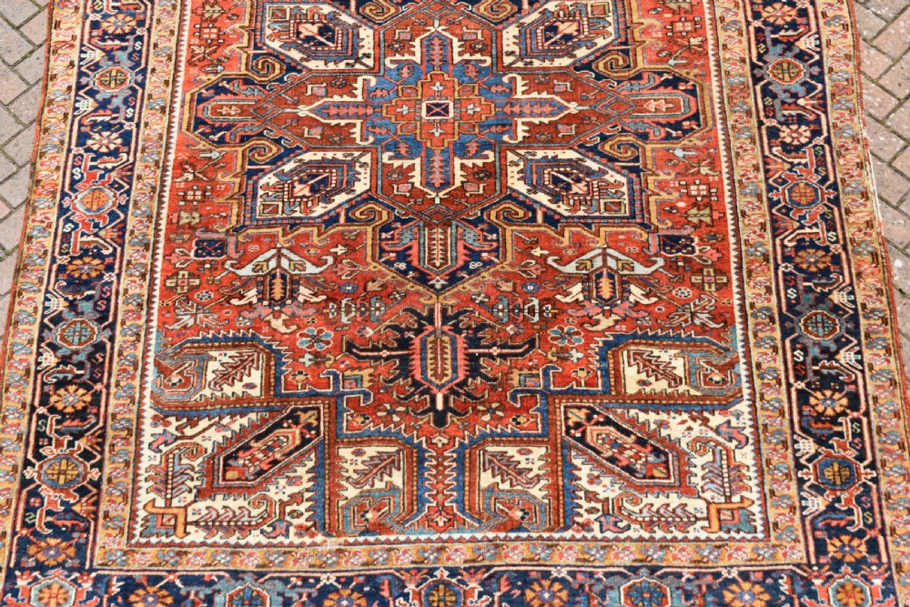 A large antique Heriz room-size carpet displaying a prominent geometric central medallion in navy blue, terracotta red, and cream tones. View II